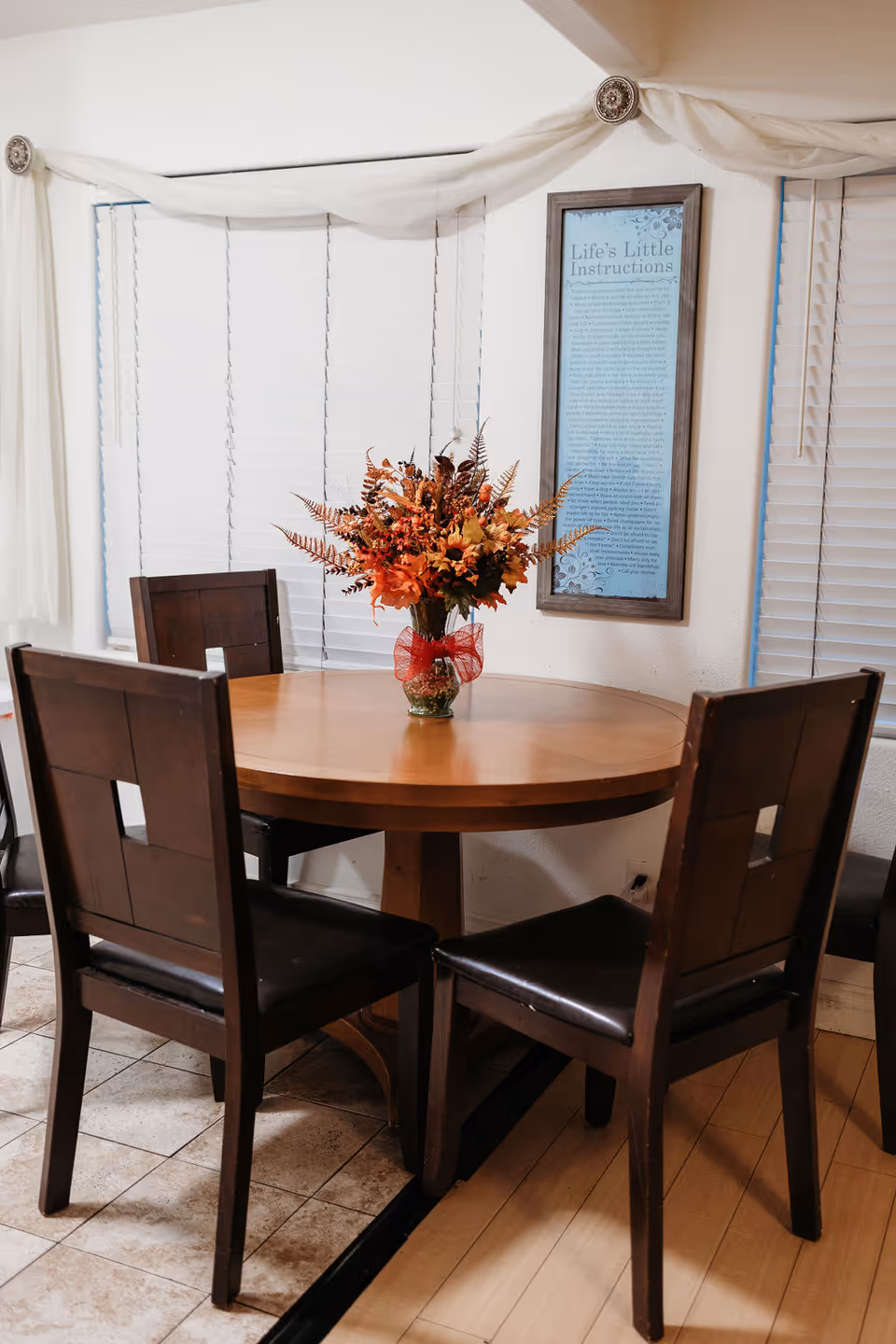 Round wooden dining table with a floral centerpiece and four chairs in a dining area with window blinds and framed wall art.