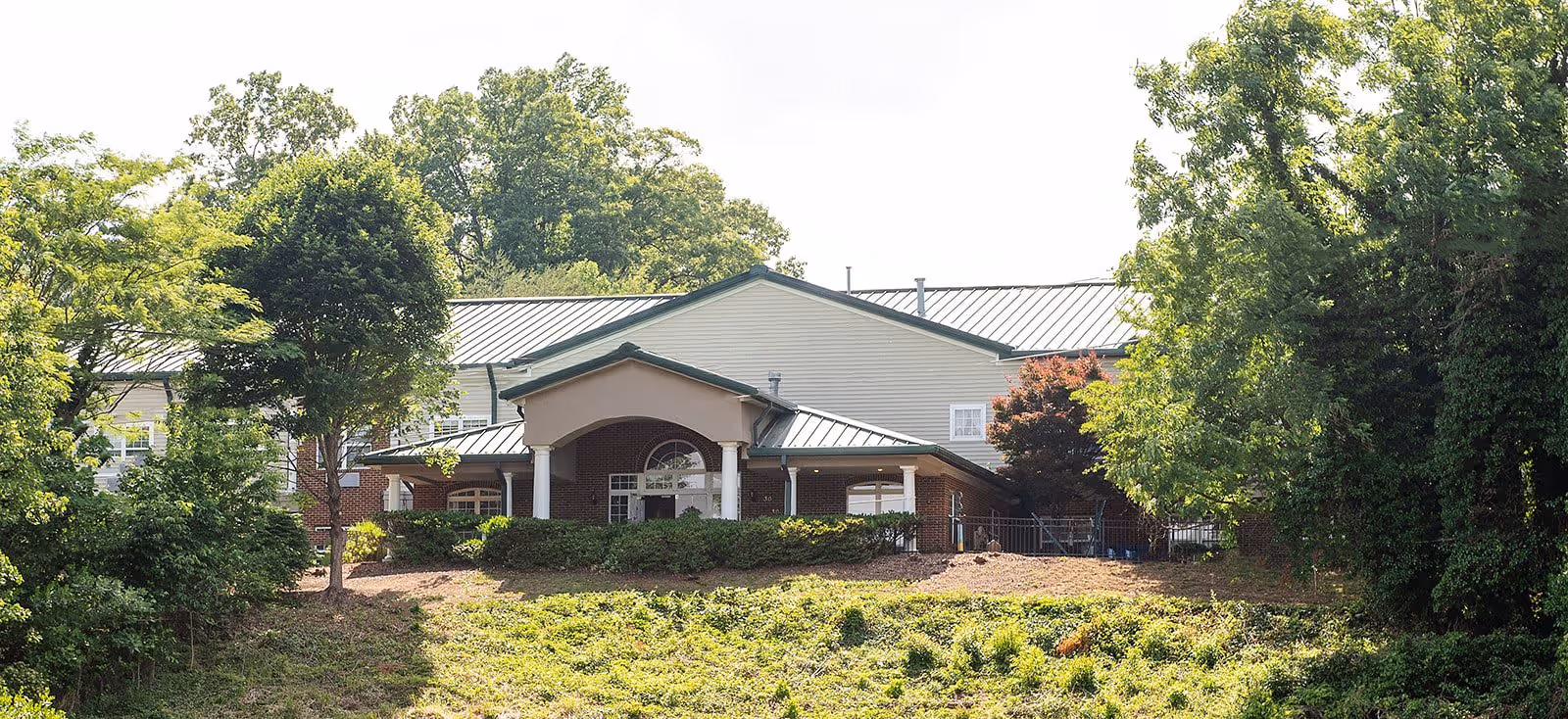 Exterior view of a senior living facility building partially obscured by trees and greenery, featuring a covered entrance with columns and a metal roof.