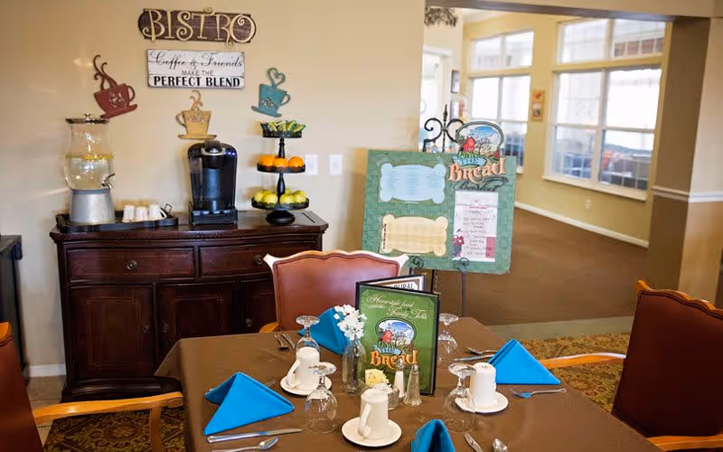 A cozy dining area in a senior living facility with a table set for four, featuring blue folded napkins, cups, glasses, and silverware. Behind the table is a wooden sideboard with a coffee maker, water dispenser, and a tiered fruit stand. Wall decor includes a sign that says 'Bistro Coffee & Friends Make the Perfect Blend.' A menu board is displayed on an easel near large windows letting in natural light.