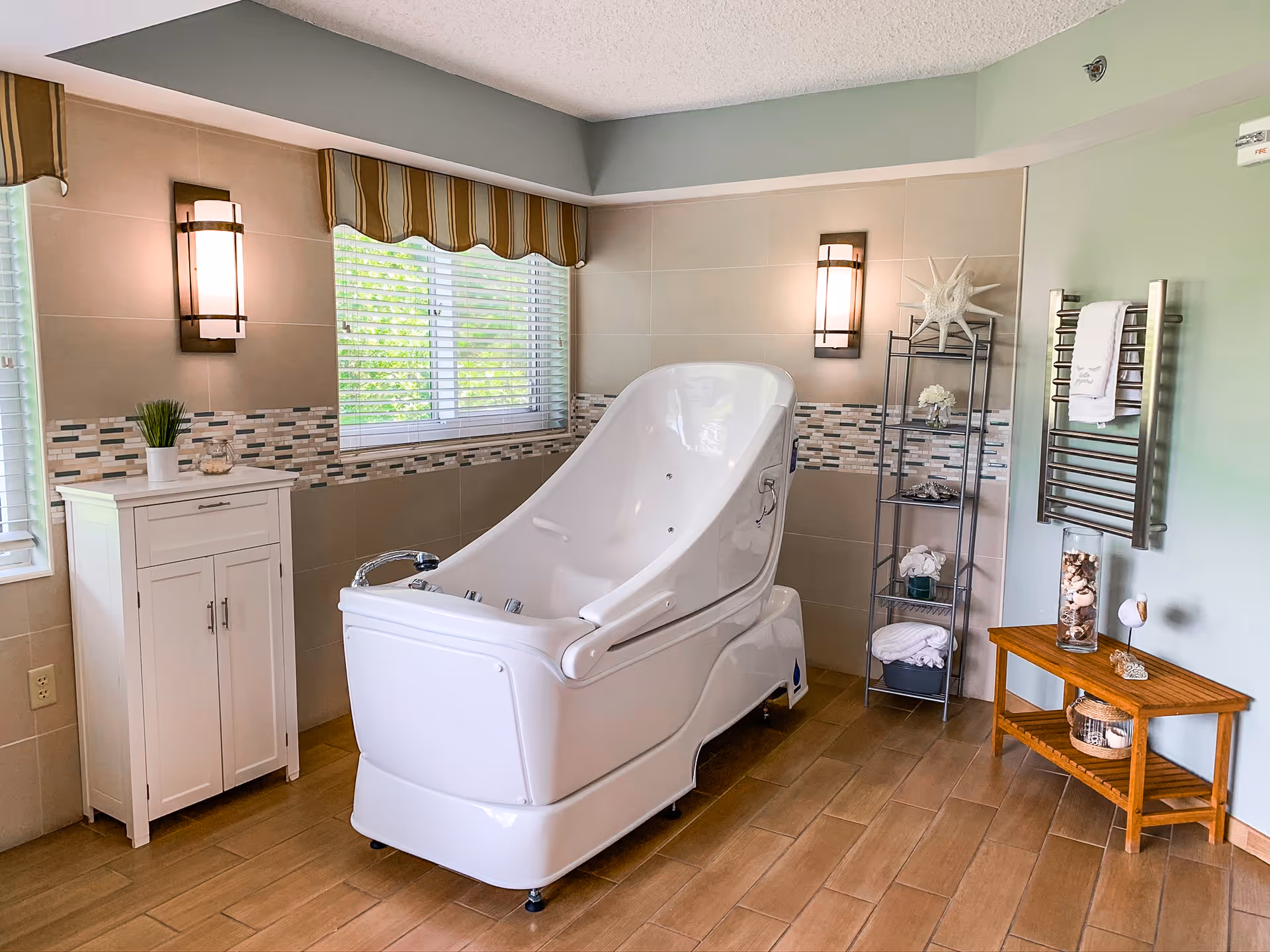 A bright and clean bathroom featuring a large white therapeutic bathtub in the center. The room has beige tiled walls with a decorative mosaic tile strip, two wall-mounted lights, a window with blinds and a striped valance, a white cabinet with a small plant and jar on top, a metal shelving unit with towels and decorative items, a wooden bench with seashell decorations, and a towel warmer mounted on the wall.