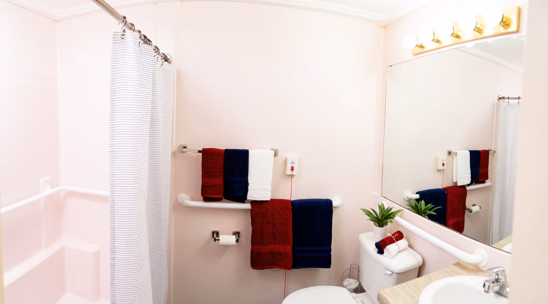 A clean and well-lit bathroom with a white shower curtain, grab bars on the walls, a toilet with folded towels on top, a sink with a countertop, a large mirror above the sink, and multiple red, white, and blue towels hanging on towel racks.
