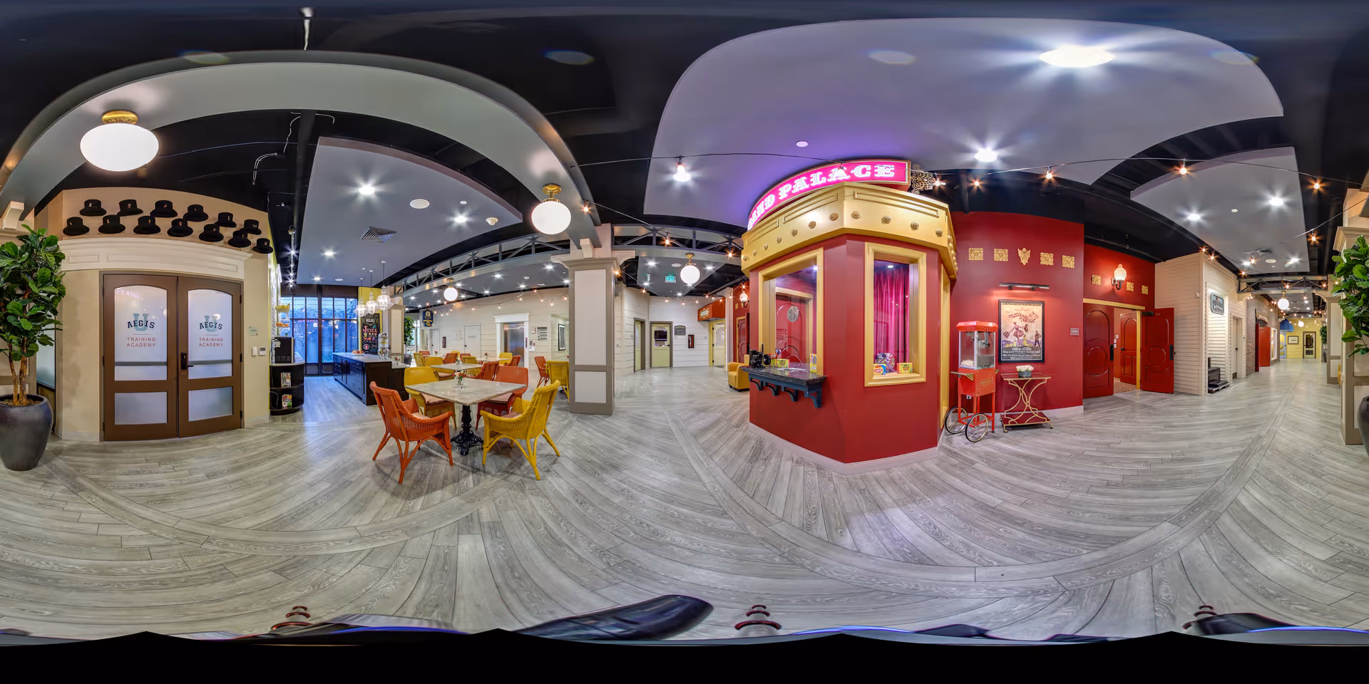 A spacious and brightly lit interior common area of Aegis Living Queen Anne Rodgers Park featuring a vintage-style popcorn stand with a neon sign, colorful chairs around tables, decorative plants, and doors labeled with the facility's name. The flooring is light wood, and the ceiling has modern lighting fixtures.