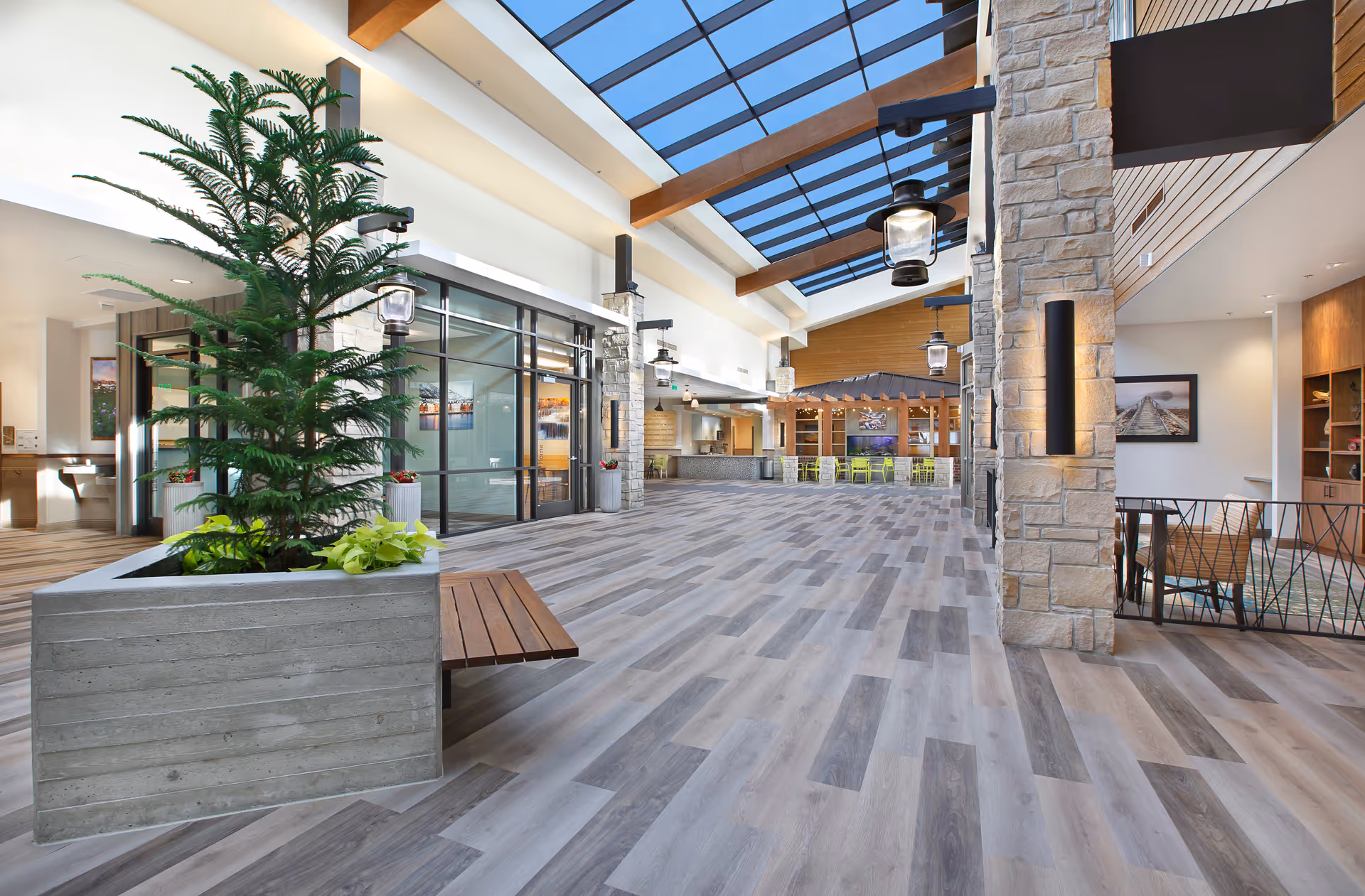 Spacious, modern indoor common area with large skylights, wood and stone accents, a planter with greenery and a built-in bench, and seating area with tables and chairs in the background.