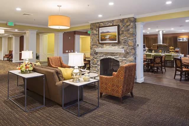 A cozy senior living facility common area featuring a stone fireplace with a framed landscape painting above it. The seating area includes a brown sofa with yellow pillows, two patterned armchairs, and side tables with lamps and decorative items. In the background, there is a dining area with tables and chairs, and a kitchen with wooden cabinets and stainless steel appliances.