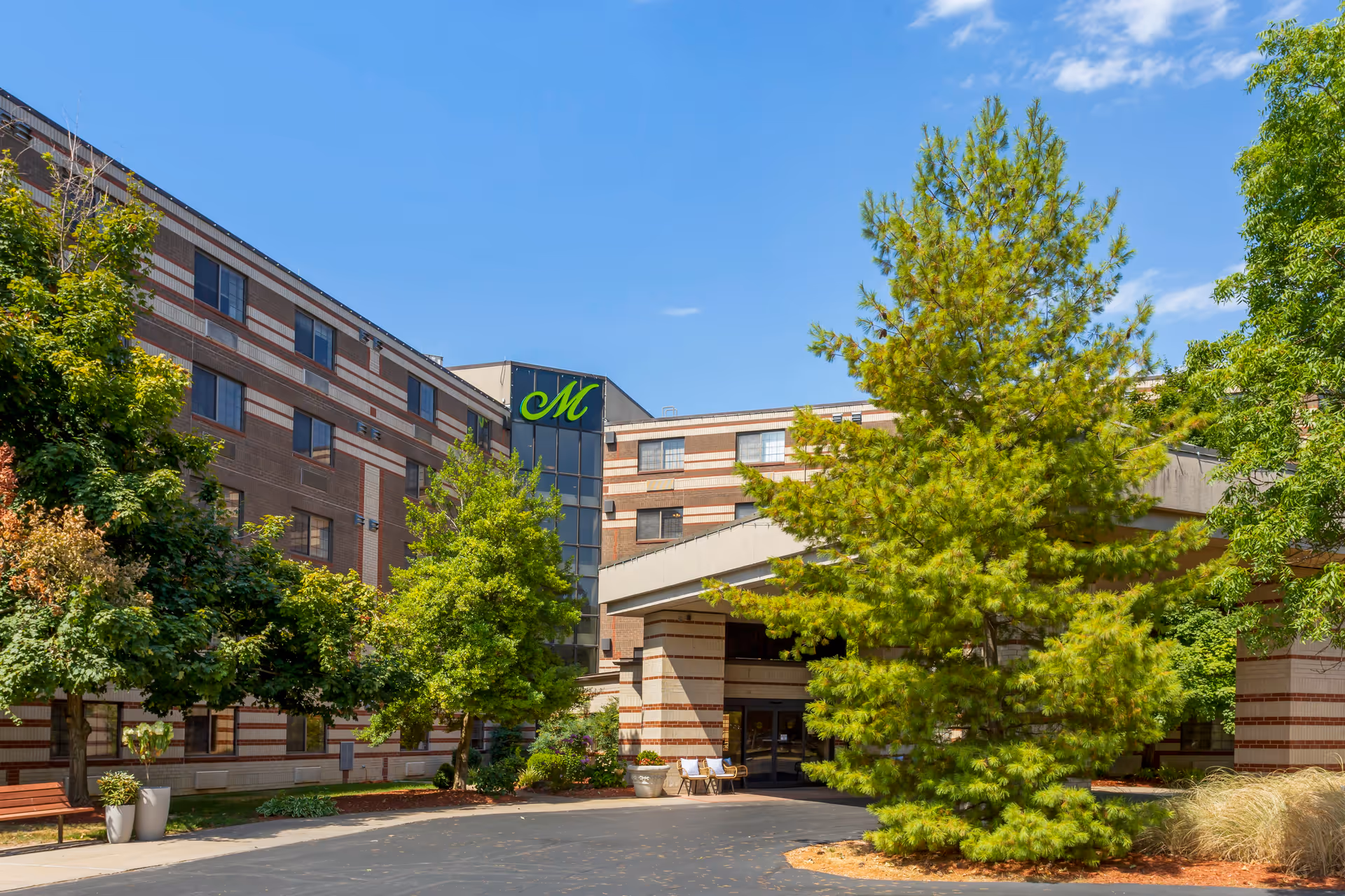 Exterior view of The Montclair Senior Living facility showing a multi-story brick building with a covered entrance, surrounded by green trees and landscaping under a clear blue sky.