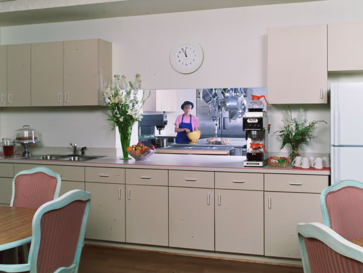 Interior view of a kitchen area with beige cabinets and drawers, a sink, a coffee maker, a bowl of fruit, and flower vases on the countertop. A window opening shows a person in a pink shirt and blue apron holding a yellow bowl in a commercial kitchen. There are pink cushioned chairs around a wooden table in the foreground.