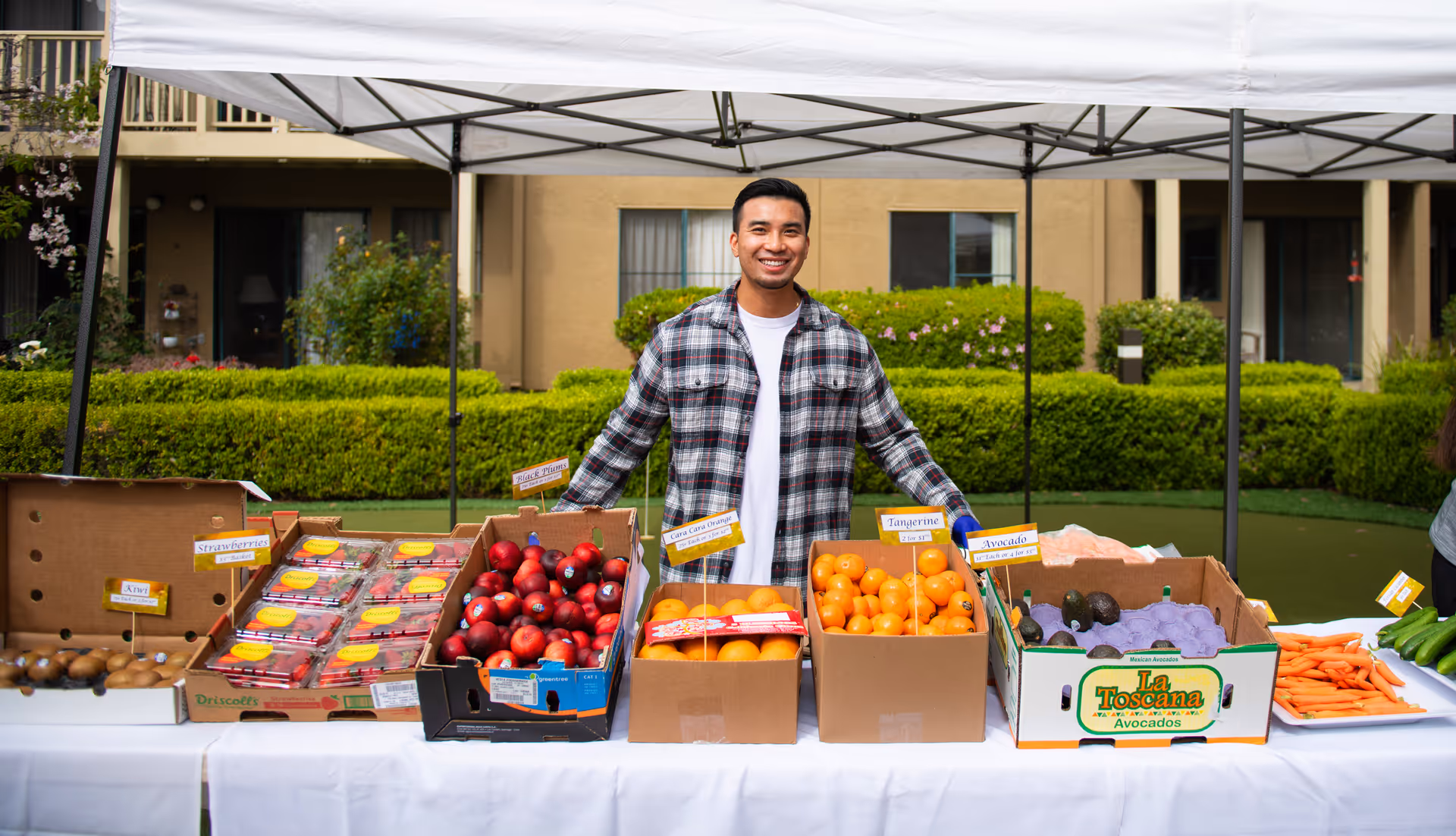 A man wearing a plaid shirt stands behind a table covered with a white cloth displaying various fresh fruits and vegetables including kiwis, strawberries, apples, oranges, tangerines, avocados, carrots, and cucumbers under a white canopy outdoors with a building and greenery in the background.