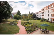 Outdoor courtyard area with a red brick walking path winding through green grass and landscaped garden beds. There are trees and shrubs along the path, and a patio area with a table, chairs, and a green umbrella. Surrounding the courtyard are multi-story residential buildings under a partly cloudy sky.