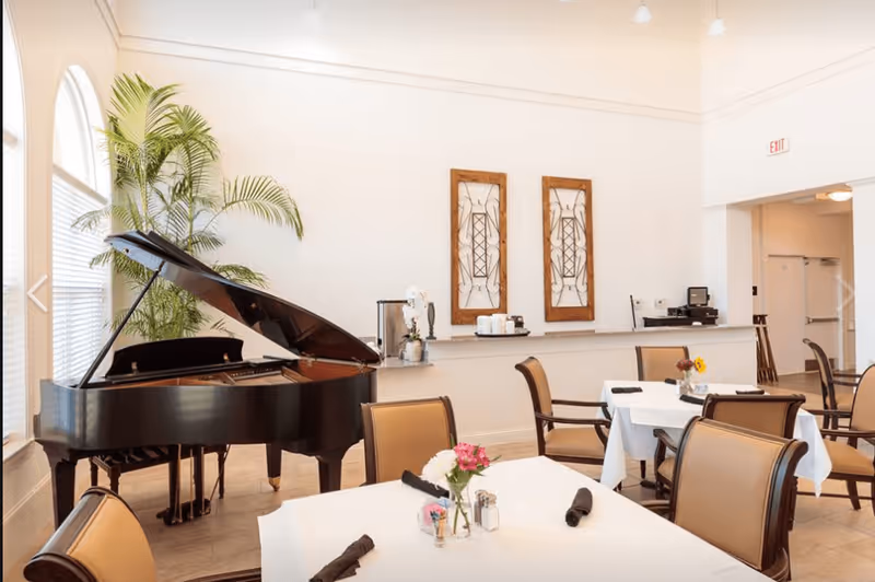 A bright dining room with tables covered in white tablecloths, each set with napkins and small flower arrangements. A black grand piano is positioned near large windows with blinds, and a tall green plant is beside it. The walls are white with two decorative wooden framed artworks. Chairs with tan cushions surround the tables.