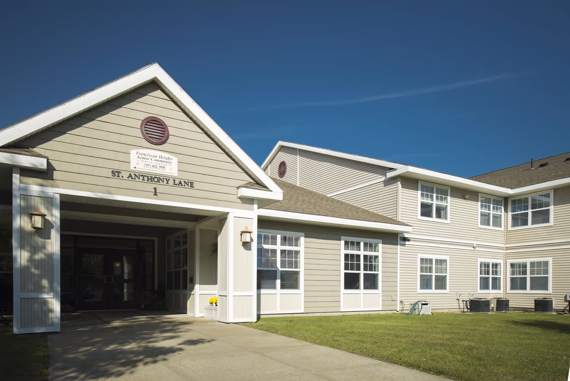 A beige, multi-wing senior living building with a covered entrance and a green lawn under a clear blue sky.