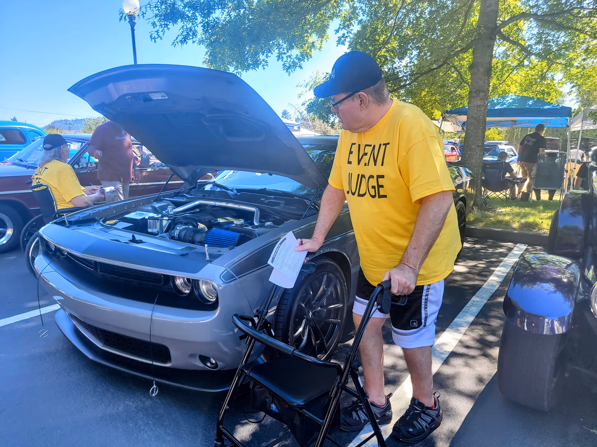 An older man wearing a yellow 'EVENT JUDGE' t-shirt and black cap is inspecting a silver car with its hood open at an outdoor car show. He is using a walker and holding a piece of paper. In the background, another person in a yellow 'EVENT JUDGE' t-shirt is sitting near a vintage car, and several people are gathered under a canopy among trees.