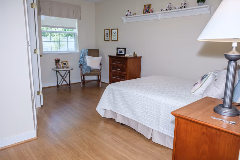 A cozy bedroom with a neatly made bed covered in a white quilt. There is a wooden nightstand with a lamp on the right side of the bed. Across from the bed, there is a wooden dresser with framed photos and decorative items on top. A comfortable chair with a pillow and a blue throw blanket is placed near a window with a beige valance. A small round table next to the chair holds a flower vase and a framed photo. The room has light-colored walls and wood flooring.