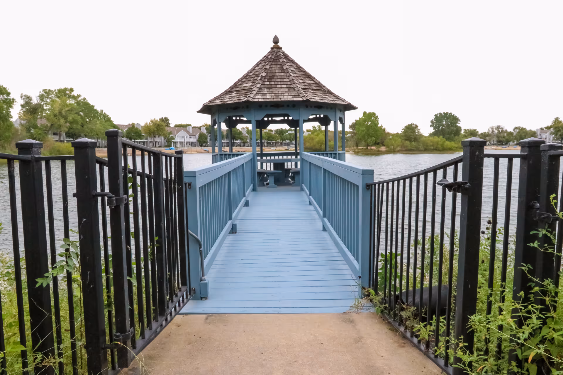 A blue wooden gazebo at the end of a blue wooden walkway extending over a body of water, with black metal gates at the entrance and green trees in the background.