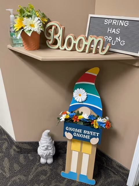 A corner of an indoor space decorated with a small shelf holding a potted plant with yellow and white flowers, a wooden sign that says 'Bloom', and a letter board with the message 'SPRING HAS PRUNG'. Below the shelf is a colorful wooden gnome figure holding a sign that reads 'GNOME SWEET GNOME', surrounded by artificial flowers, and a small white garden gnome statue on the floor next to it.