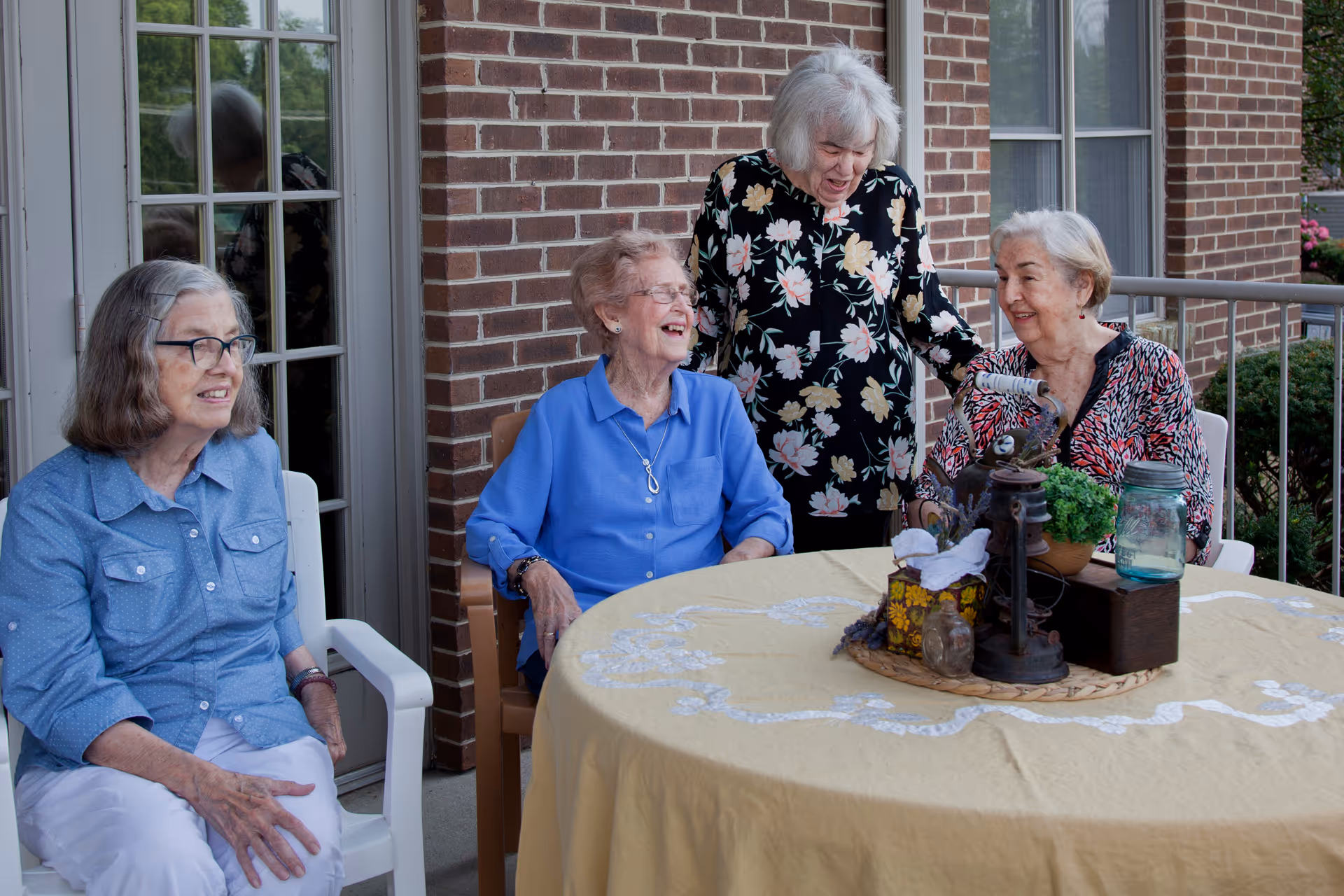 Four elderly women sitting and standing around a round table with a yellow tablecloth on a patio outside a brick building. The table has decorative items including a lantern, a small plant, and a jar. The women are smiling and appear to be enjoying each other's company.