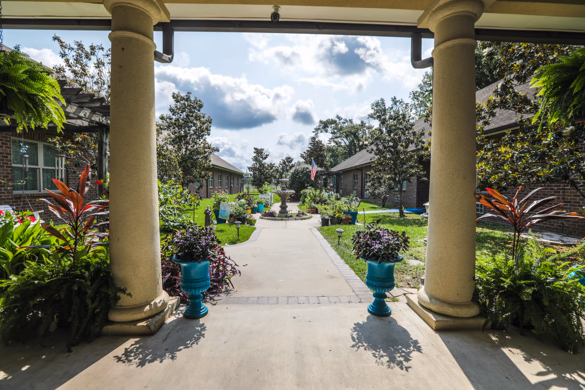 View through a covered patio with two large columns looking out onto a landscaped garden path with potted plants, trees, and a central fountain. Brick buildings with windows flank the garden on both sides under a partly cloudy sky.