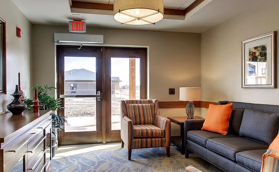 A cozy seating area in a senior living facility with a striped armchair, a dark gray sofa with orange pillows, a wooden side table with a lamp, a wooden cabinet with decorative vases, and glass double doors leading outside. The room has beige walls, a patterned carpet, and a ceiling light fixture.