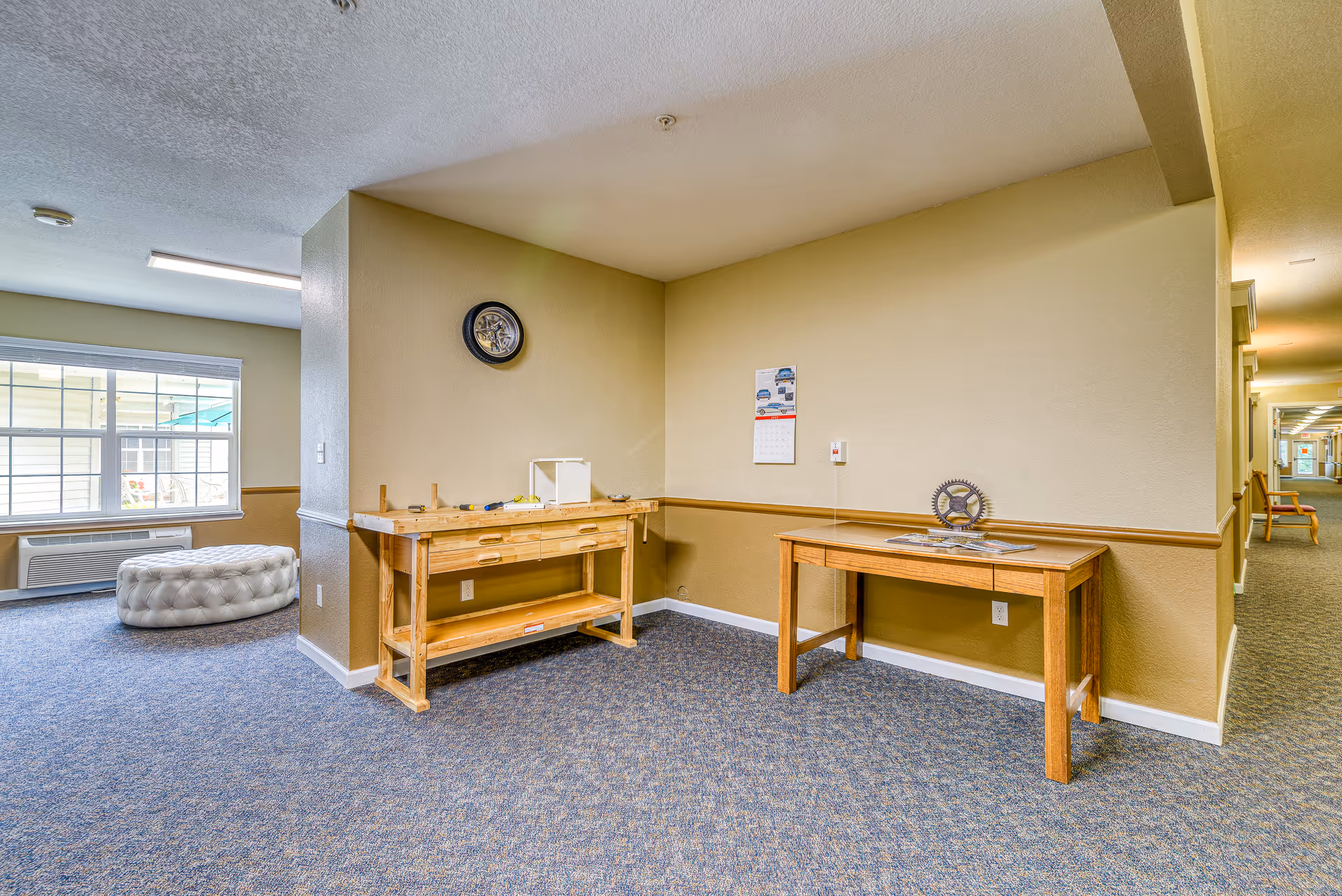 Interior corner of a memory care facility with two wooden tables against beige walls. One table has tools and a small white box, while the other has a decorative gear and some magazines. A clock and a calendar hang on the walls. Carpeted floor and a hallway with chairs are visible in the background.