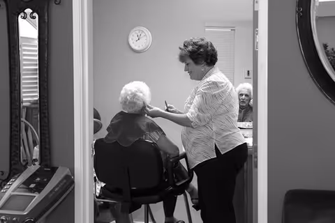 A woman is styling the hair of an elderly woman seated in a chair inside a room. Another elderly woman is visible in the background, sitting and looking towards the camera. The room has a clock on the wall and a mirror reflecting part of the scene.