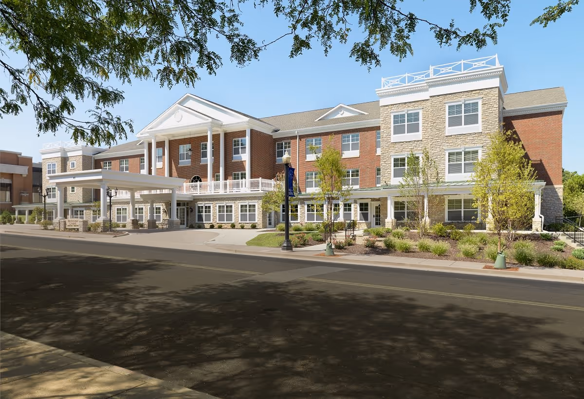 Front exterior of a three-story brick and stone senior living facility with a covered porte-cochère, landscaping, and a street in the foreground.