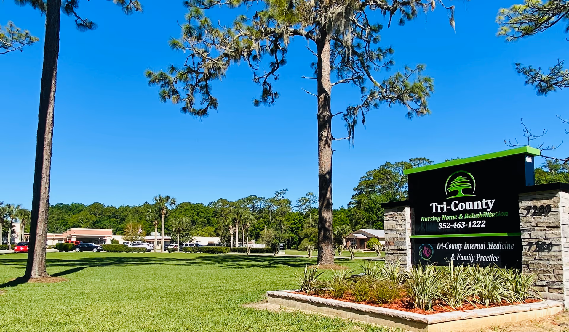 Outdoor view of the Tri-County Nursing Home facility sign with a clear blue sky, green grass, trees, and a parking area with cars in the background.