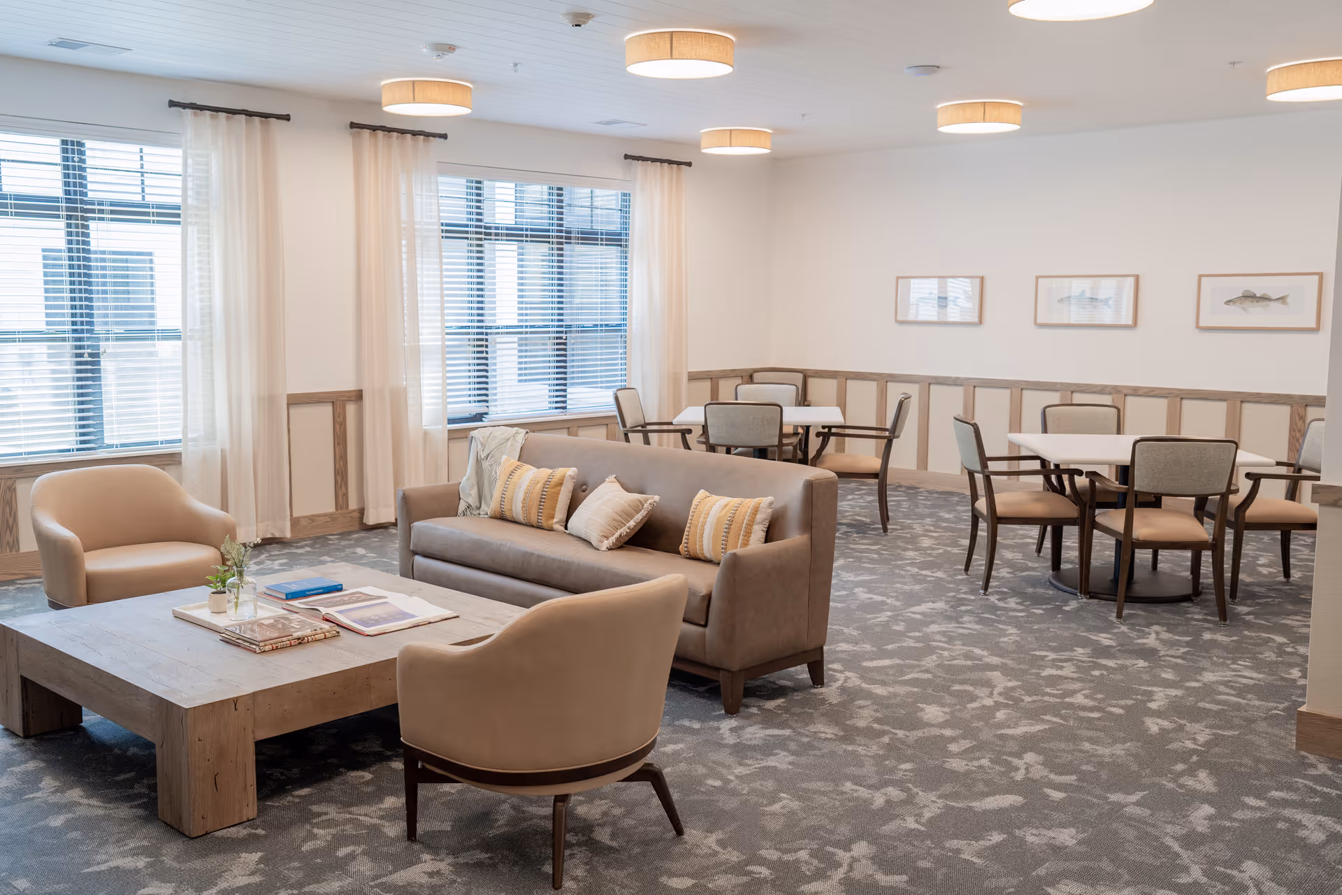 Open common room with a neutral-toned sofa, armchairs, a large coffee table and several dining tables beneath round pendant lights by large windows.