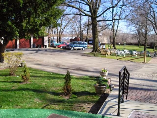 Grassy courtyard and parking area with trees, potted plants, a ramped walkway with railing, parked cars and a red outbuilding in the background.
