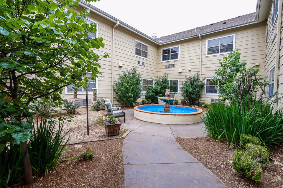A courtyard area at Aberdeen Heights Assisted Living featuring a circular water fountain in the center, surrounded by green shrubs and plants. There are beige buildings with multiple windows enclosing the courtyard, and a paved walkway leading to the fountain. Two chairs and a small planter with flowers are placed near the fountain.