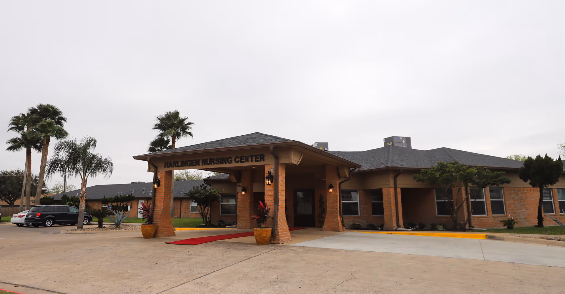 Exterior view of Harlingen Nursing Center building with a covered entrance, brick walls, and several windows. There are palm trees and other plants around the building, and a few cars parked nearby under an overcast sky.