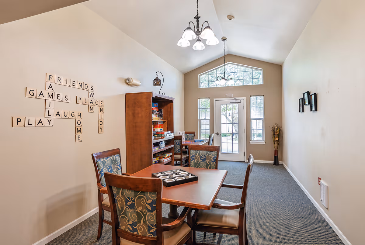 A bright game room with a wooden table and four upholstered chairs, a bookshelf filled with board games, and a wall decorated with Scrabble-style tiles spelling words like FRIENDS, GAMES, LAUGH, PLAY, FAMILY, HOME, PLACE, WIN, and ENJOY. The room has a high ceiling with two hanging light fixtures and a door with windows leading outside.