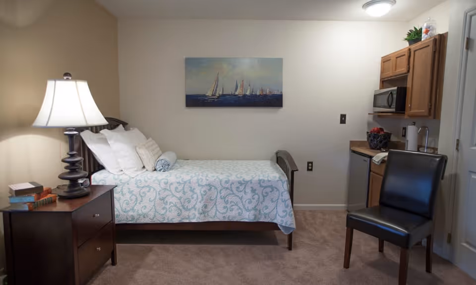 A small, cozy bedroom with a single bed covered in a light blue and white patterned quilt. The bed has several pillows and a bolster. Next to the bed is a dark wooden nightstand with a lamp and a few books. On the wall above the bed is a painting of sailboats on the water. To the right, there is a kitchenette area with wooden cabinets, a microwave, a small refrigerator, and a sink. A black leather chair is positioned near the kitchenette. The room has beige walls and carpeted flooring.