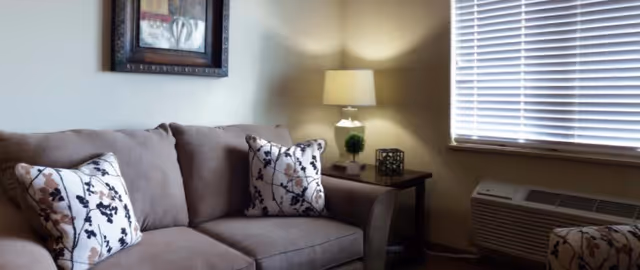 A cozy living room with a brown sofa adorned with two decorative pillows featuring a floral pattern. Next to the sofa is a wooden side table with a lamp, a small green plant, and a decorative cube. A window with closed blinds is on the right side, and below it is an air conditioning unit. A framed artwork hangs on the wall above the sofa.