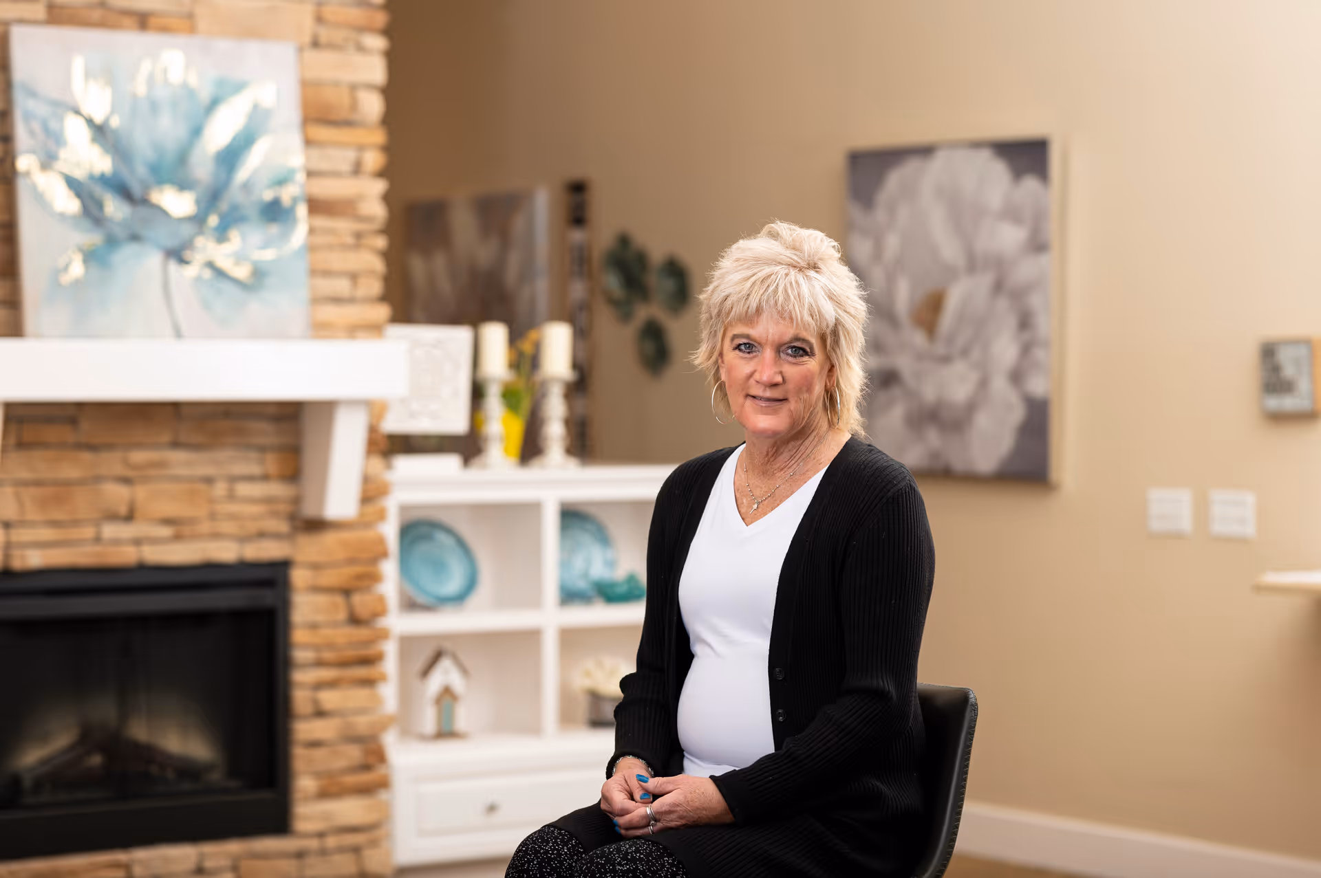 A woman with short blonde hair wearing a white shirt and black cardigan sits on a chair in a cozy living room. Behind her is a stone fireplace with a blue floral painting above it, and a white shelving unit with decorative plates and candles. The walls are beige with additional floral artwork.