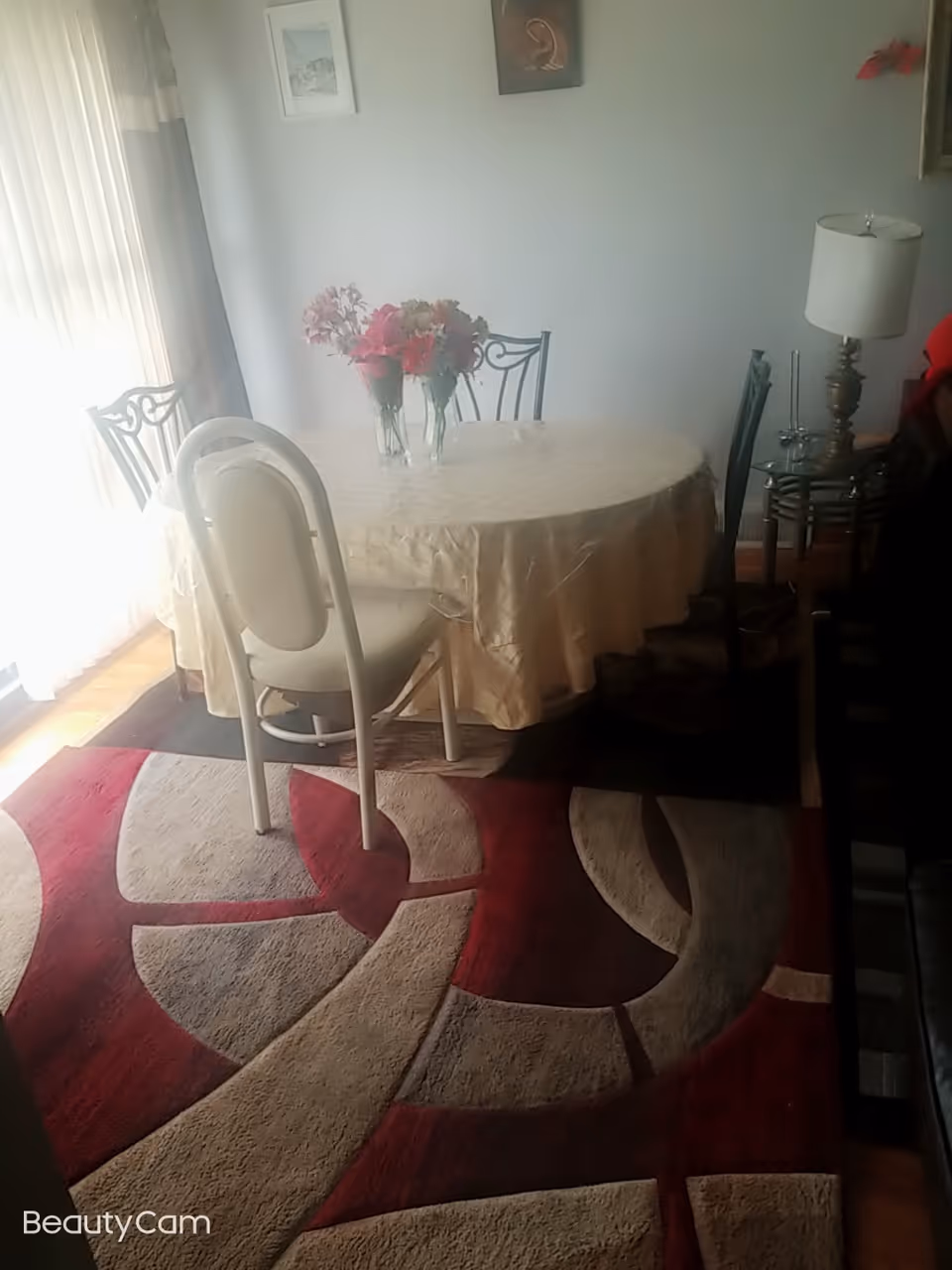 A dining area with a round table covered with a beige tablecloth, surrounded by chairs. Two vases with pink and red flowers are placed on the table. The room has a patterned red, beige, and brown rug on the floor, a lamp on a side table, and framed pictures on the wall. Light filters in through vertical blinds on the left side.