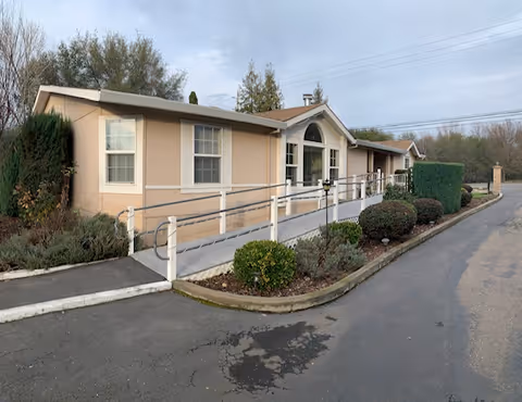 Single-story beige senior living building with ramp, shrubs, and a paved driveway, front entrance.