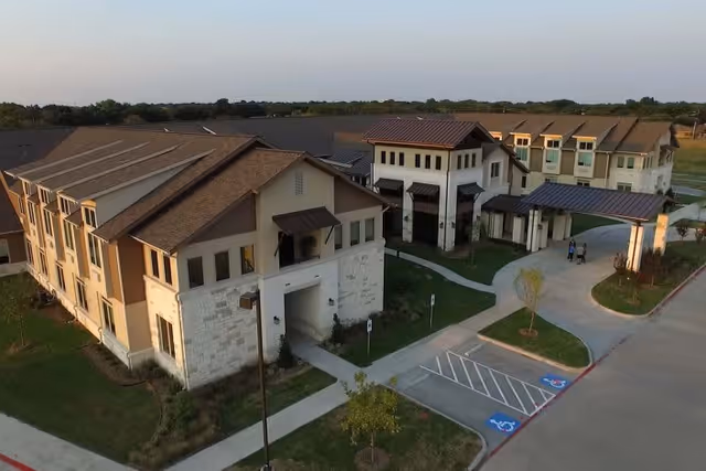 Aerial view of The Healthcare Resort of Plano showing a large, multi-story building with beige and stone exterior walls, brown roofs, and a covered entrance driveway. There are sidewalks, landscaped areas with small trees, and handicap parking spaces visible in the parking lot.