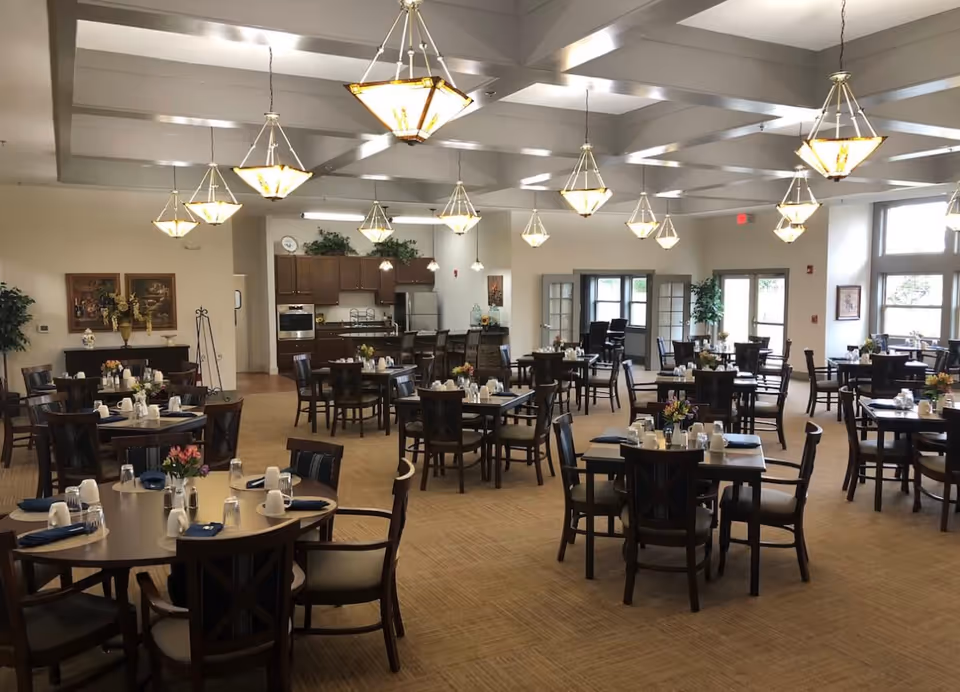 A spacious dining room with multiple dark wood tables and chairs arranged neatly. Each table is set with white cups, glasses, and napkins, and some have small flower arrangements. The room features beige carpeting, cream-colored walls, and several hanging pendant lights with warm yellow shades. In the background, there is a kitchen area with wooden cabinets, a refrigerator, and an oven. Large windows and glass doors allow natural light to enter the room.