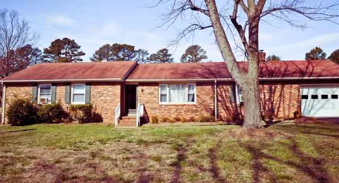 Brick single-story building with a red roof, lawn, and a large tree in front.