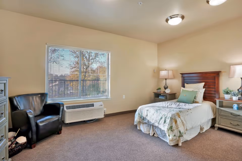 A cozy bedroom in Almond Heights featuring a single bed with white and green bedding, two bedside tables with lamps and small plants, a black leather armchair, a large window with blinds, and a beige carpeted floor.