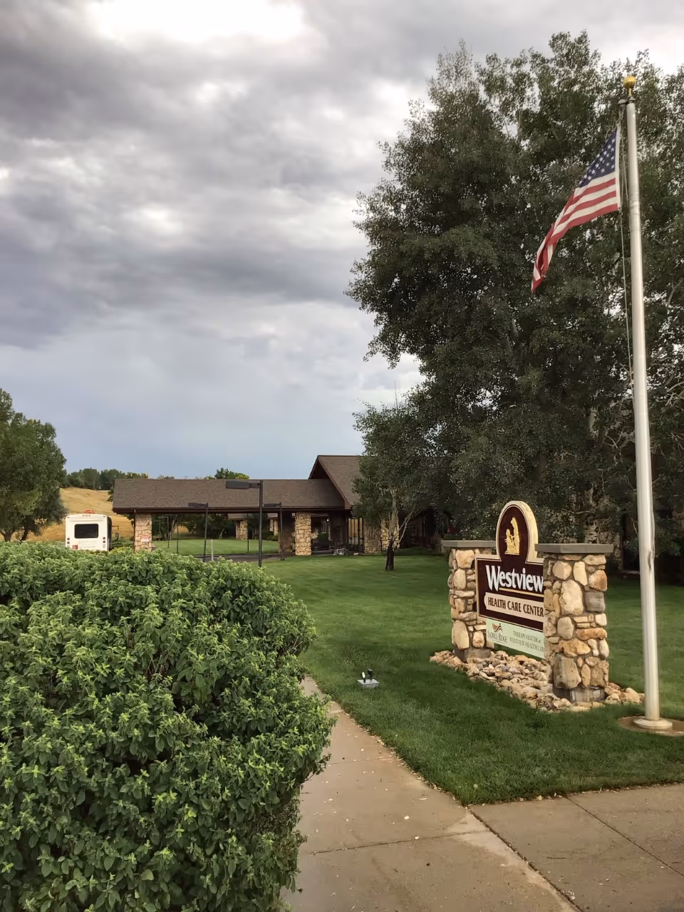Entrance and front lawn of Westview Health Care Center featuring a stone sign, an American flag on a pole, and the building under a cloudy sky.