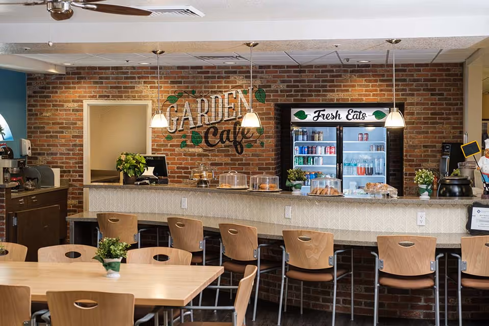 A brick-walled café counter labeled 'Garden Cafe' with a refrigerated 'Fresh Eats' display, pastries, and seating at a bar and tables.