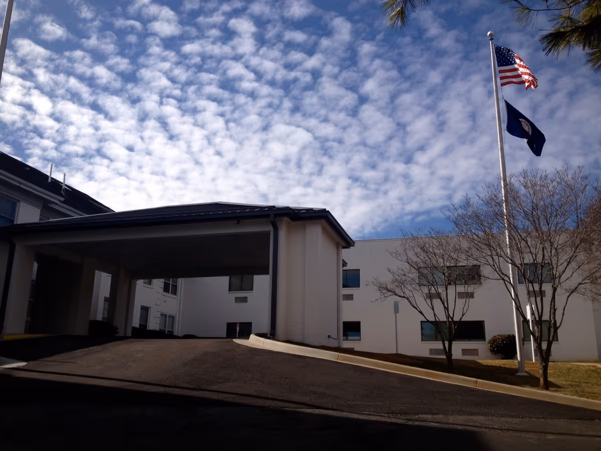 Exterior view of a building with a covered entrance driveway, two flagpoles with the American flag and another flag, and leafless trees under a partly cloudy sky.