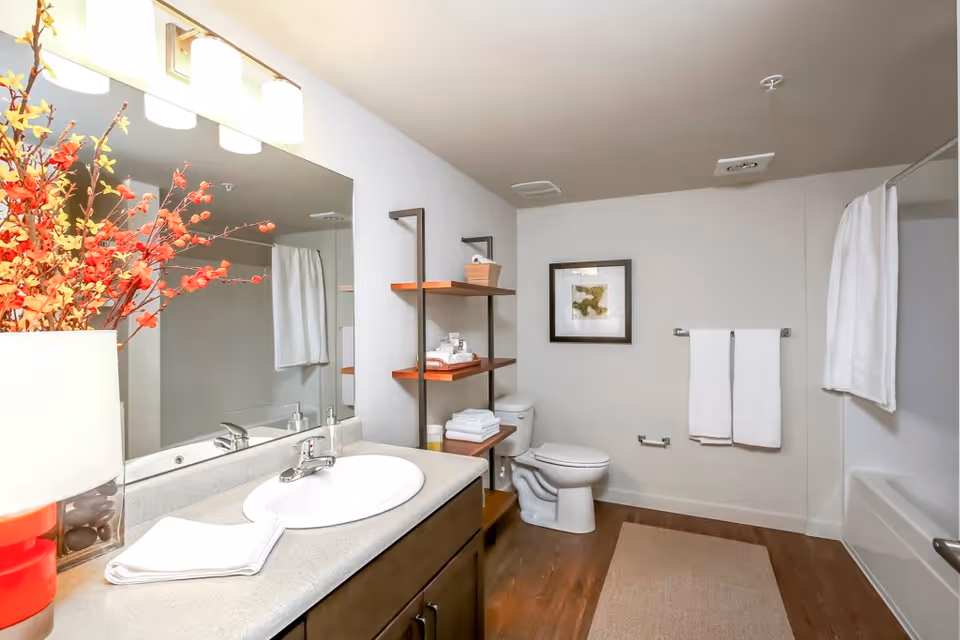 A clean and modern bathroom with a large mirror above a sink countertop. There is a vase with red and yellow flowers on the left side of the counter. The bathroom features a toilet, a bathtub with a white shower curtain, a towel rack with two white towels, and wooden shelves holding neatly folded towels and toiletries. The floor has a wooden finish with a beige rug.