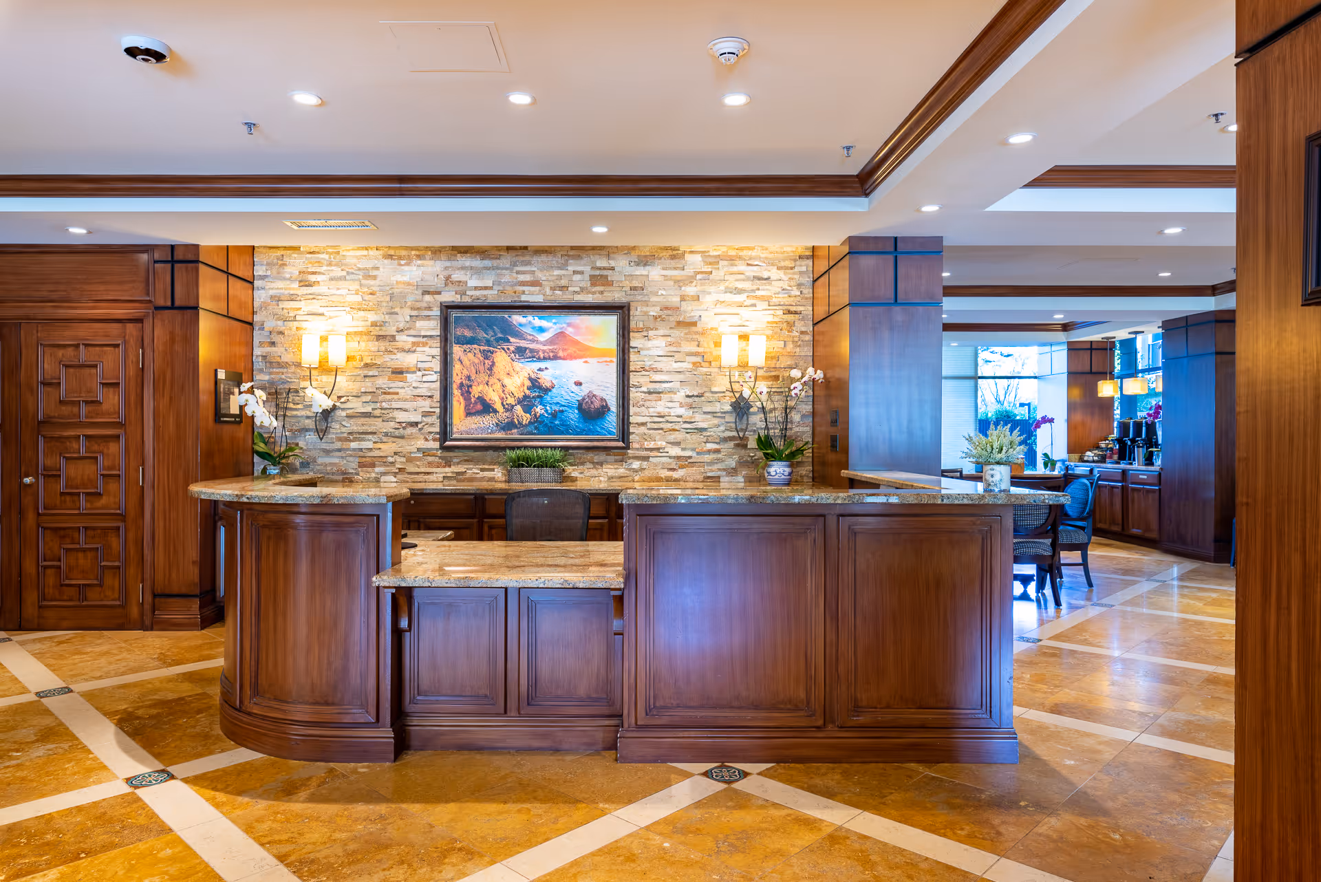 Reception desk area with wooden cabinetry and granite countertops, stone accent wall with a framed coastal landscape painting, wall sconces, and decorative plants. The floor has a polished tile design and the space is well-lit with recessed ceiling lights.