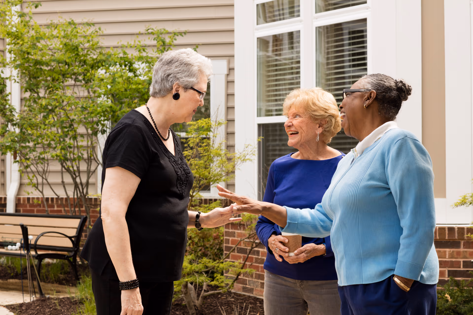Three elderly women standing outside near a building with beige siding and white-framed windows, engaged in a cheerful conversation. One woman is wearing a black shirt, another a blue sweater, and the third a light blue cardigan. There is a bench and some greenery in the background.