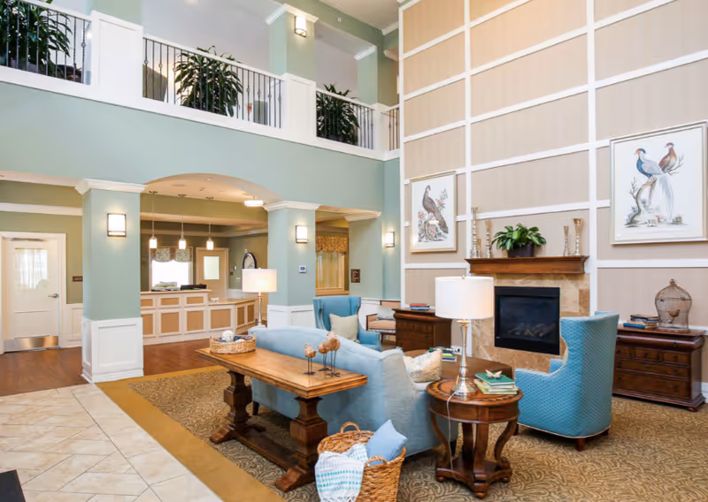 Bright two-story senior living lobby with seating area, fireplace, and a reception desk in the background.