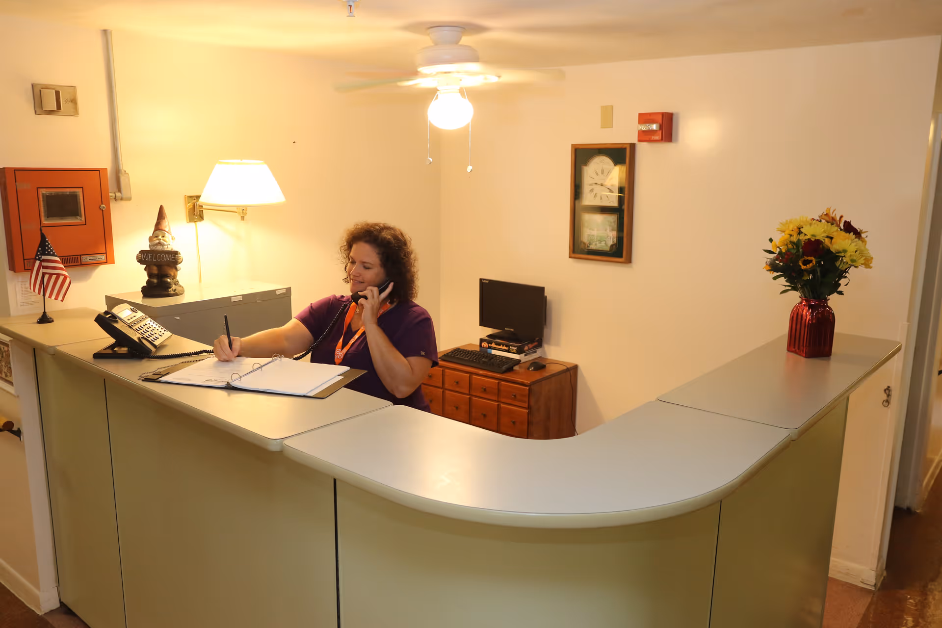 A woman sitting behind a reception desk in a facility, talking on the phone and writing in a binder. The desk has a small American flag, a lamp, a 'Welcome' gnome, and a vase with flowers. There is a small computer and a framed picture on the wall behind her.