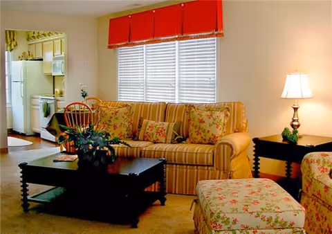 A cozy living room with a striped sofa adorned with floral cushions, a matching floral ottoman, a dark wooden coffee table with a plant on it, and a side table with a lamp. In the background, there is a kitchen area with white appliances and cabinetry.
