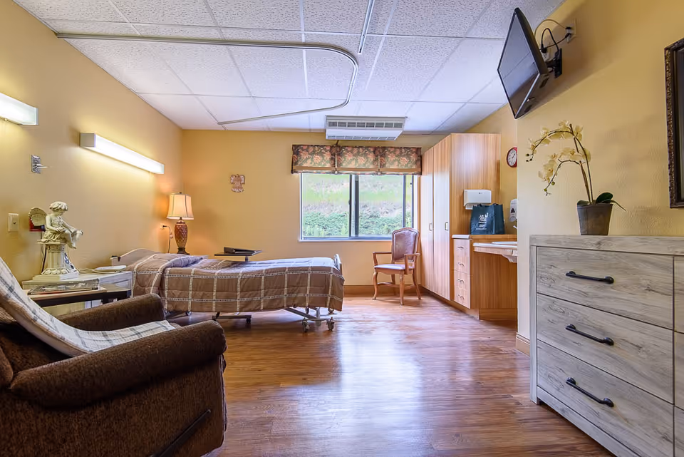 A well-lit senior living facility bedroom with a single bed covered in a brown checkered blanket, a reclining chair with a blanket draped over it, a wooden dresser with a potted orchid, a wall-mounted TV, a wooden chair near a window with floral valance, and built-in wooden cabinets and countertop.