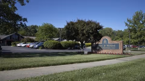 Exterior view of Arbors at Milford senior living facility showing a parking lot with several cars, trees, bushes, and a sign with the facility name near a sidewalk.